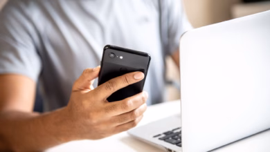 Mann hält vor einem laptop sein smartphone in der hand